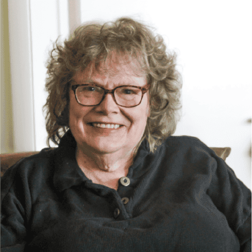 An older woman with curly gray hair and glasses smiles while sitting indoors, wearing a dark button-up shirt.