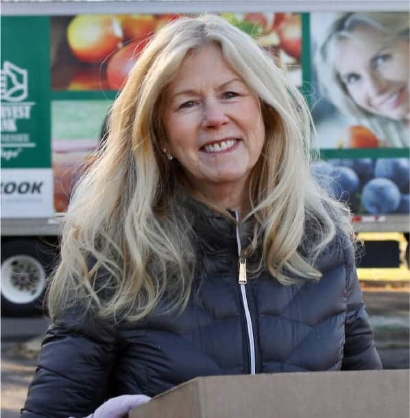 A smiling woman with long blonde hair, wearing a dark jacket, stands outdoors holding a box. Behind her is a truck with colorful fruit images and a womans face displayed on its side.