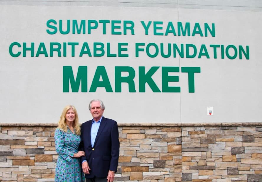 A man and woman stand together, smiling, in front of a building with a stone facade and a sign that reads Sumpter Yeaman Charitable Foundation Market.