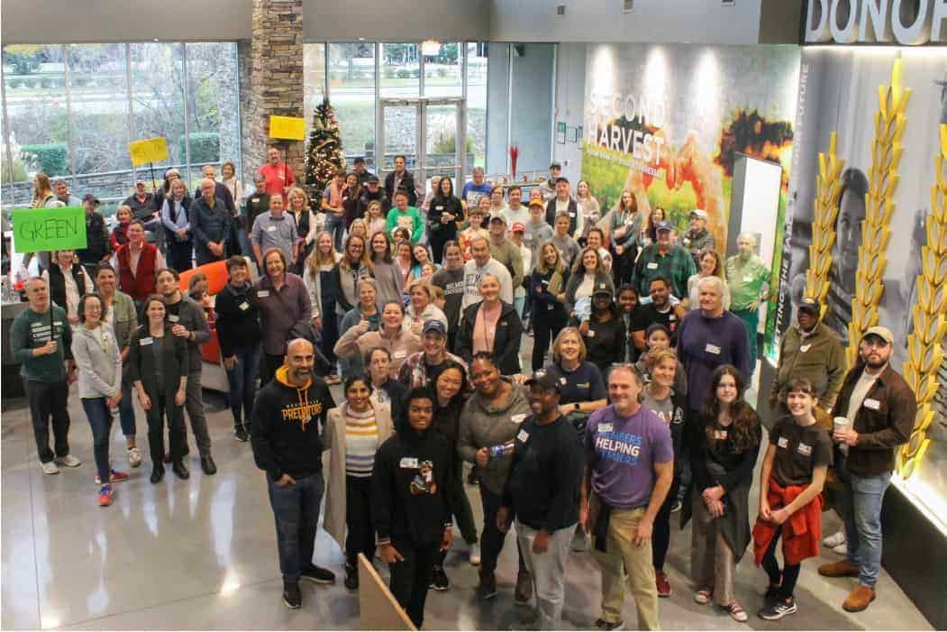 A large group of diverse people gathers indoors, many smiling and posing for a group photo. Some hold colorful signs, and there’s a decorated Christmas tree and a wall with the word “HARVEST” in the background.