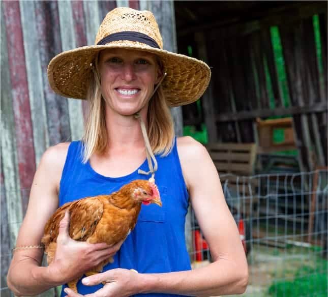 A smiling person wearing a wide-brimmed straw hat and a blue sleeveless top holds a brown chicken in front of a rustic barn and wire fence.