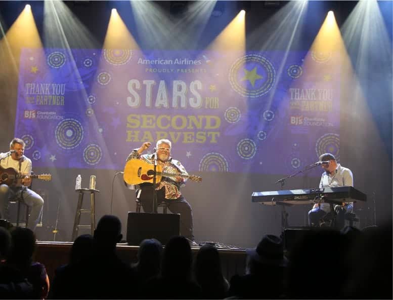 Three musicians perform on stage with guitars and a keyboard under spotlights. Behind them is a large screen displaying “Stars for Second Harvest” and sponsorship logos. Audience silhouettes are visible in the foreground.