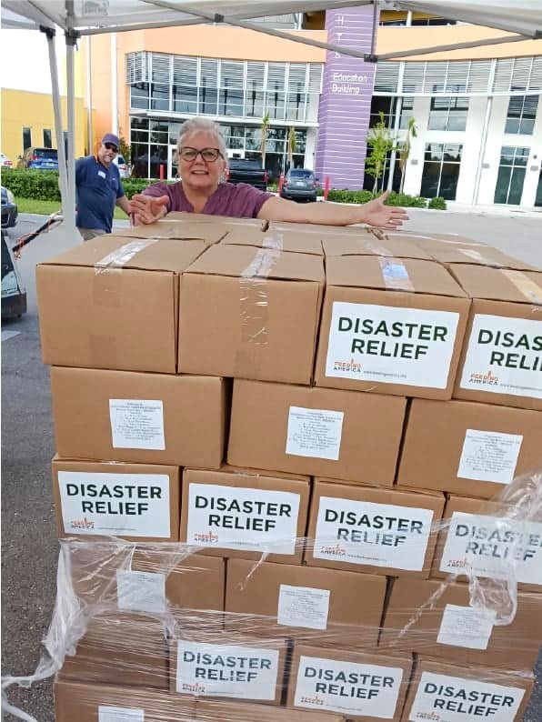 An older woman smiles behind a large stack of boxes labeled DISASTER RELIEF outside a building, while a man stands in the background. The boxes are shrink-wrapped and ready for distribution.