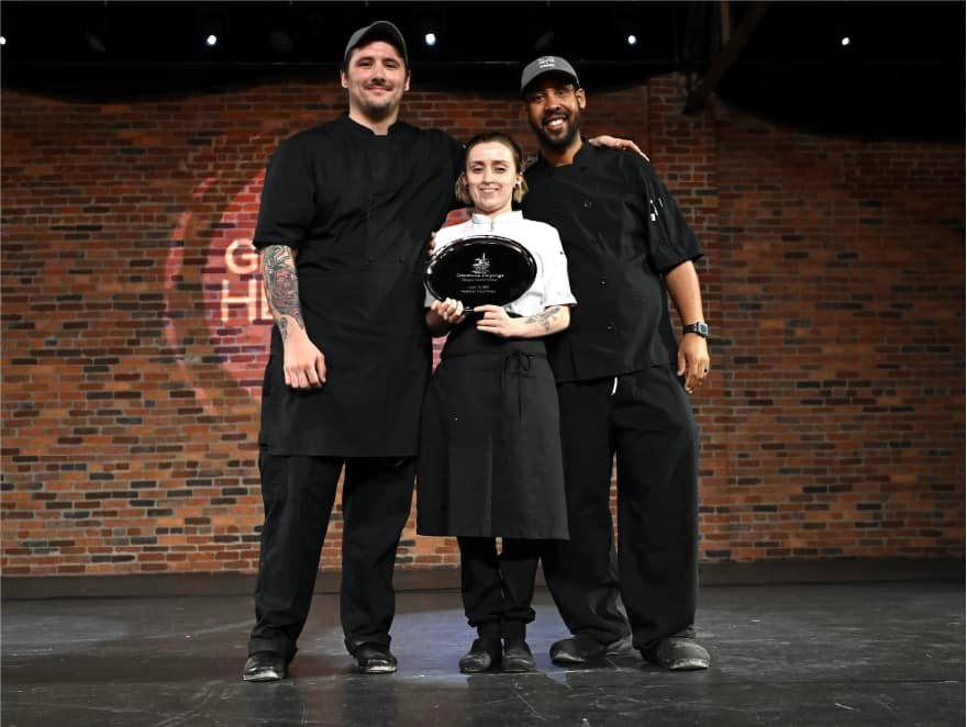 Three chefs in black uniforms stand on stage in front of a brick wall. The chef in the center holds a black award plate, while the two others stand on either side with arms around her shoulders, all smiling.