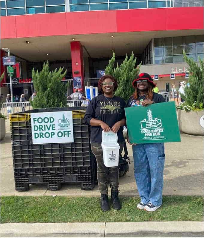 Two people stand smiling outside a building near large bins labeled FOOD DRIVE DROP OFF, holding containers and a sign for Second Harvest Food Bank, encouraging food donations.