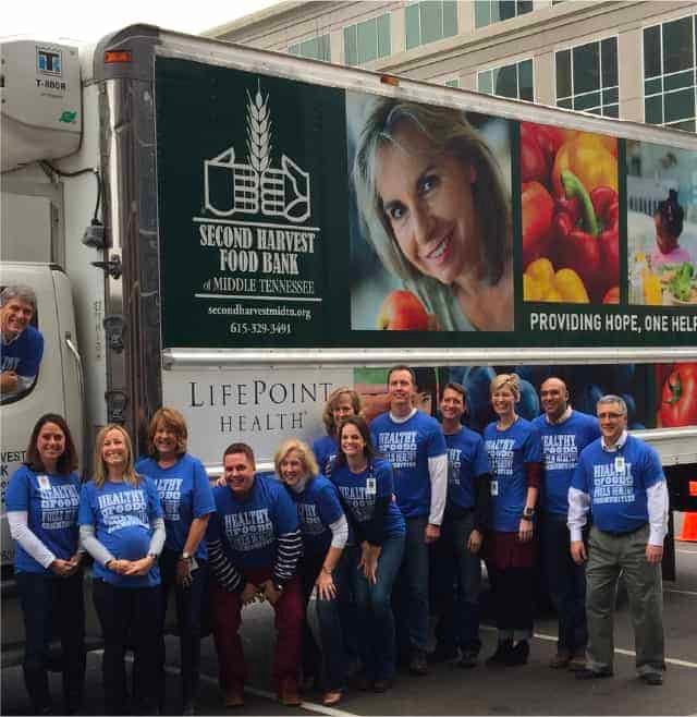 A group of people in matching blue shirts stands smiling in front of a Second Harvest Food Bank truck parked outside an office building.