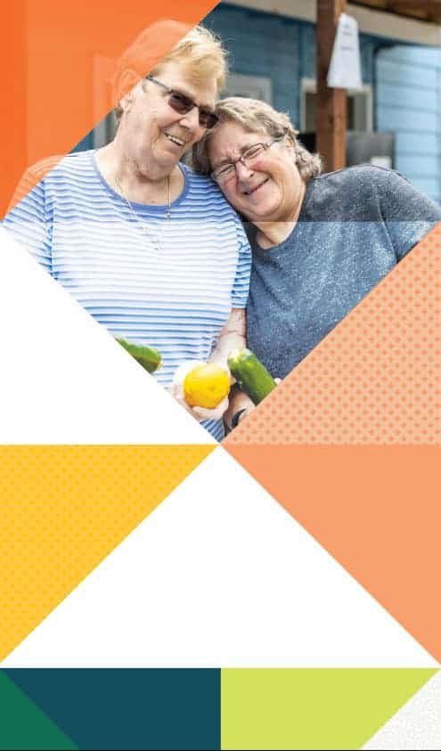 Two smiling older women stand close together outdoors, each holding garden vegetables. Colorful geometric shapes frame the image, with orange, yellow, green, and white triangular patterns at the edges.