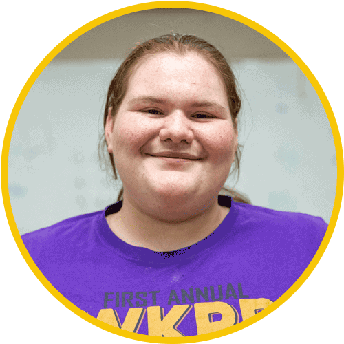A person with fair skin and long hair smiles at the camera, wearing a purple T-shirt with yellow lettering. The photo is framed by a yellow circular border.