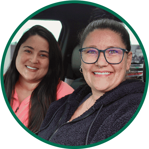 Two women smiling while sitting in a car. One wears glasses and a dark zip-up jacket; the other has long dark hair and a bright pink top. The photo is framed in a green circle.