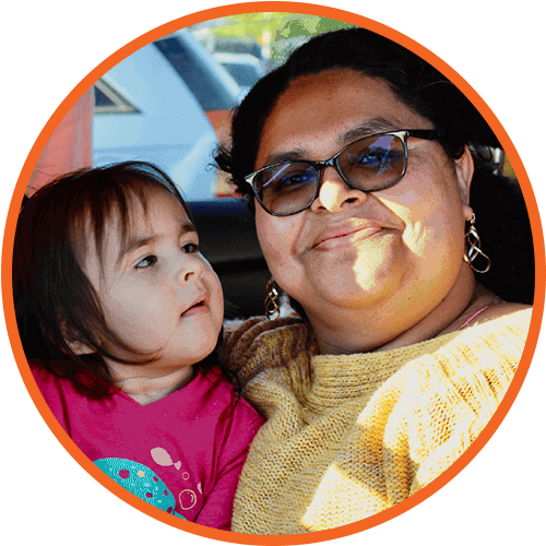 A smiling woman in glasses and a yellow sweater holds a young child in a pink shirt. The child looks up at the woman. The image is cropped in a circle with an orange background.