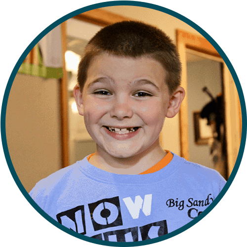 Smiling young boy with short brown hair wearing a light blue shirt, standing indoors with a missing front tooth visible.