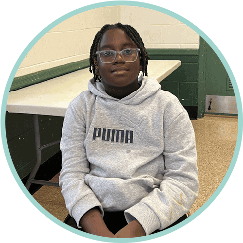 A young person with glasses and braided hair sits indoors, wearing a gray Puma hoodie and smiling slightly. A folded table and a green-and-white wall are visible in the background.
