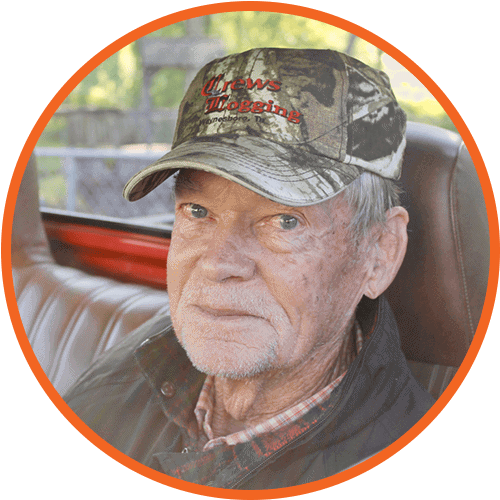 An older man with light skin, blue eyes, and a weathered face sits in a vehicle. He wears a camouflage cap with red lettering, a black jacket, and a plaid shirt. The image has an orange circular border.