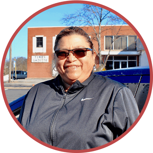 A person wearing sunglasses and a black Nike jacket smiles while standing outdoors in front of a brick building labeled Times Gazette. A blue car and leafless tree branches are visible in the background.