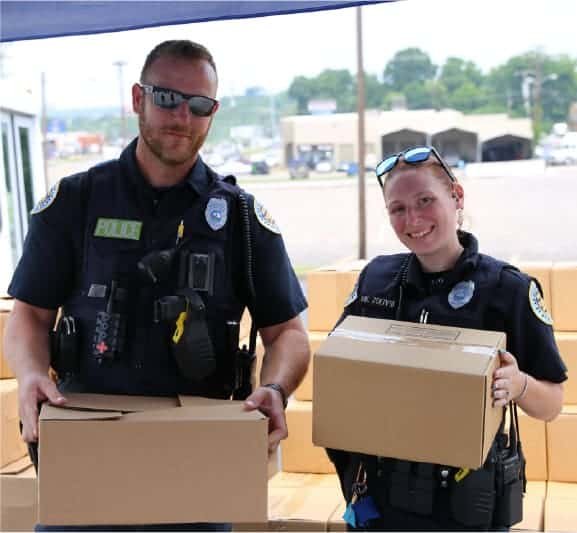 Two police officers, one man and one woman, wearing uniforms and sunglasses, smile while holding cardboard boxes outdoors, standing in front of a table and some buildings in the background.