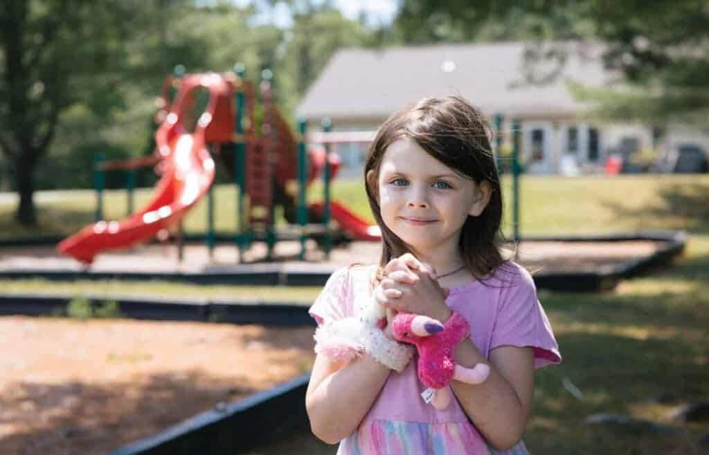 A young girl with brown hair in a pink dress stands outside, holding a pink stuffed animal, with a playground and red slide in the background on a sunny day.