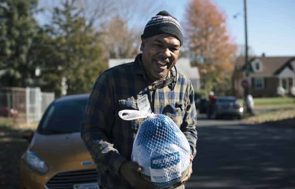 A smiling man in a plaid shirt and knit hat stands outdoors holding a frozen turkey in a net bag, with houses, trees, and a yellow car in the background on a sunny day.