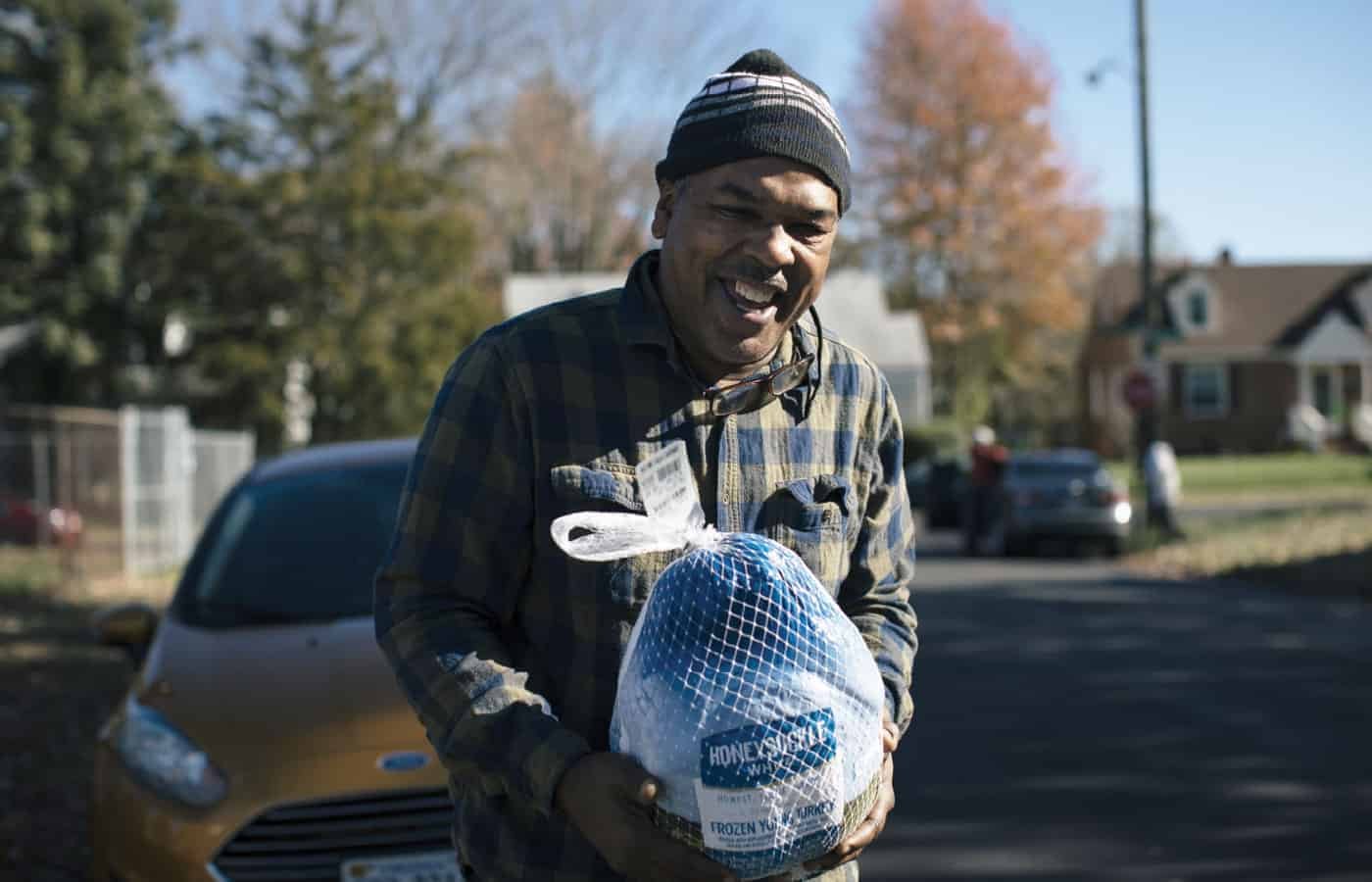 A smiling man in a plaid shirt and knit hat stands outdoors holding a frozen turkey in a net bag, with houses, trees, and a yellow car in the background on a sunny day.