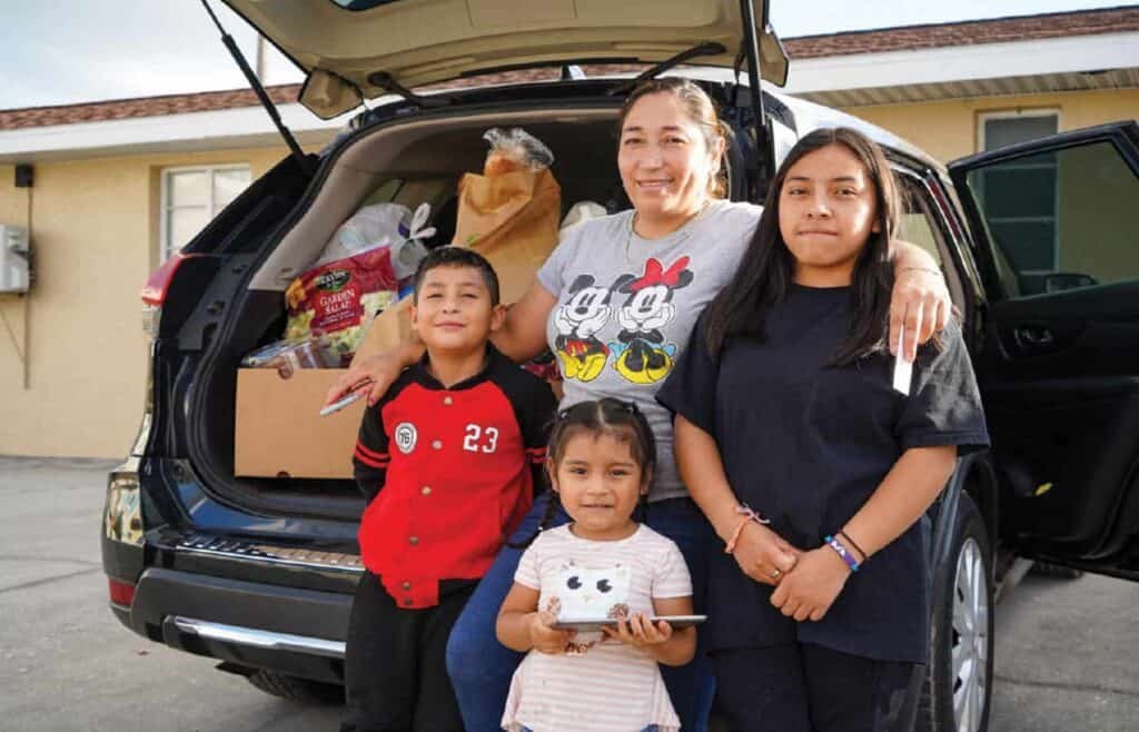 A smiling woman with three children stands in front of an open car trunk filled with groceries and supplies, posing together outside a building on a sunny day.