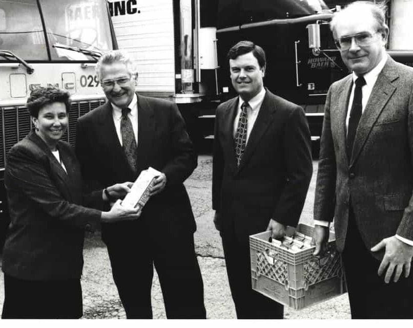 Four adults in business attire stand outdoors near trucks. One woman hands a packaged item to a smiling man while another man holds a crate of cans. All appear to be engaged in a formal or ceremonial presentation.