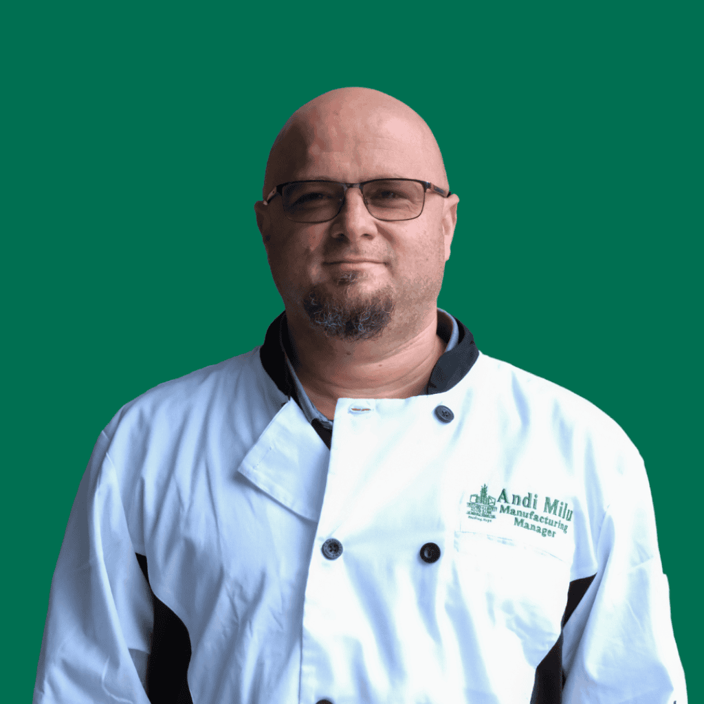 A man with glasses and a goatee wearing a white chef coat with black trim stands against a solid green background. The coat reads “Andi Milly, Manufacturing Manager.”.