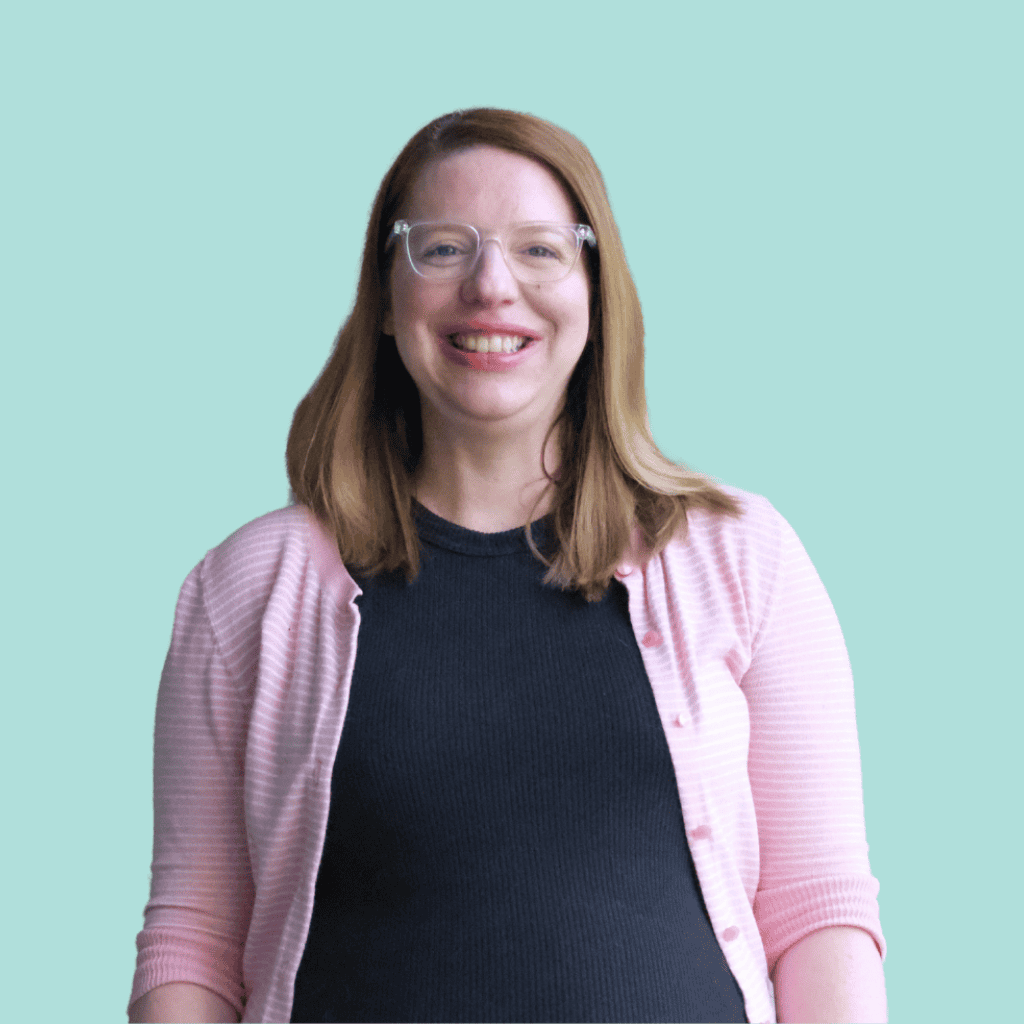 A smiling woman with straight, shoulder-length light brown hair wears clear glasses, a black top, and a light pink cardigan, standing in front of a plain light blue background.