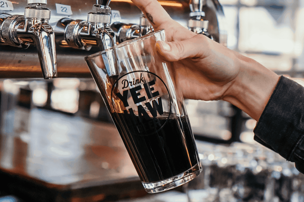 A hand holds a glass with the words “Drink Yee Haw” while filling it with dark beer from a tap at a bar.
