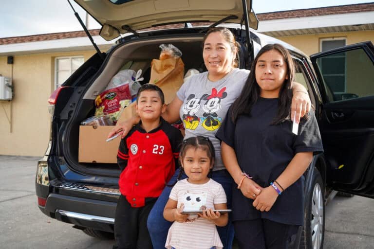 A woman and three children smile in front of an open SUV trunk filled with groceries and household items, standing outside a house on a sunny day.