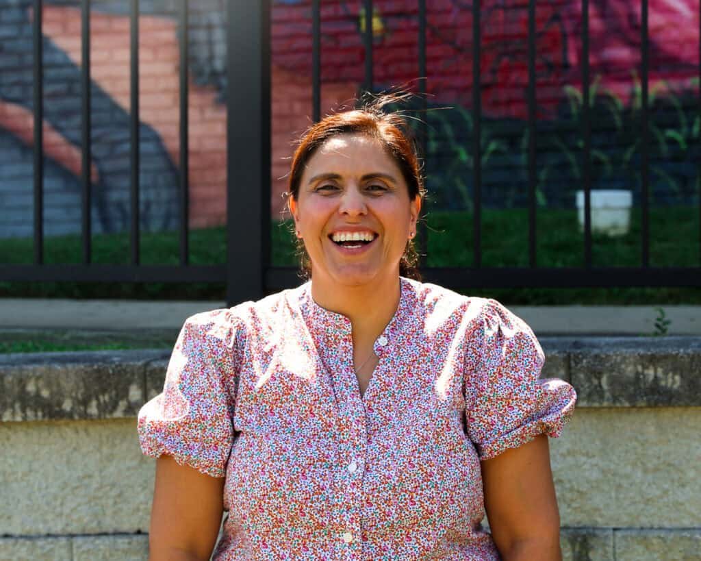 A woman with dark hair tied back, wearing a short-sleeved, floral blouse, sits outside and smiles brightly. There is a fence and a brick wall with graffiti in the background.