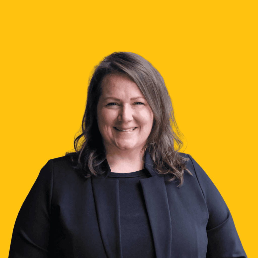A woman with wavy brown hair, wearing a black blazer, smiles in front of a solid yellow background.