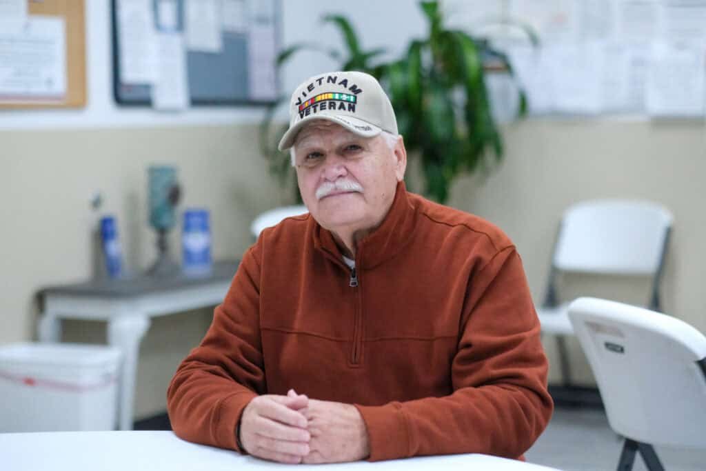 An older man with a mustache, wearing a tan Vietnam Veteran cap and an orange-brown zip-up sweater, sits at a table in a room with white chairs and bulletin boards in the background.