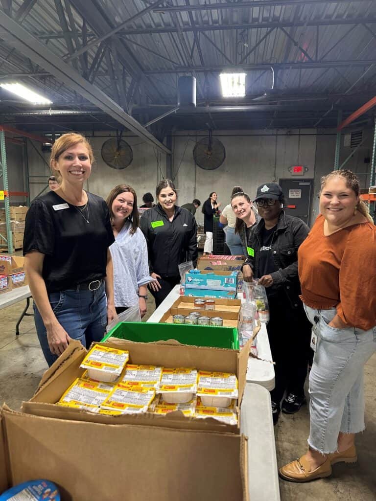 A group of six smiling people stand behind a table packed with food items, such as noodles and snacks, inside a warehouse. They appear to be volunteering or participating in a food drive or packaging event.