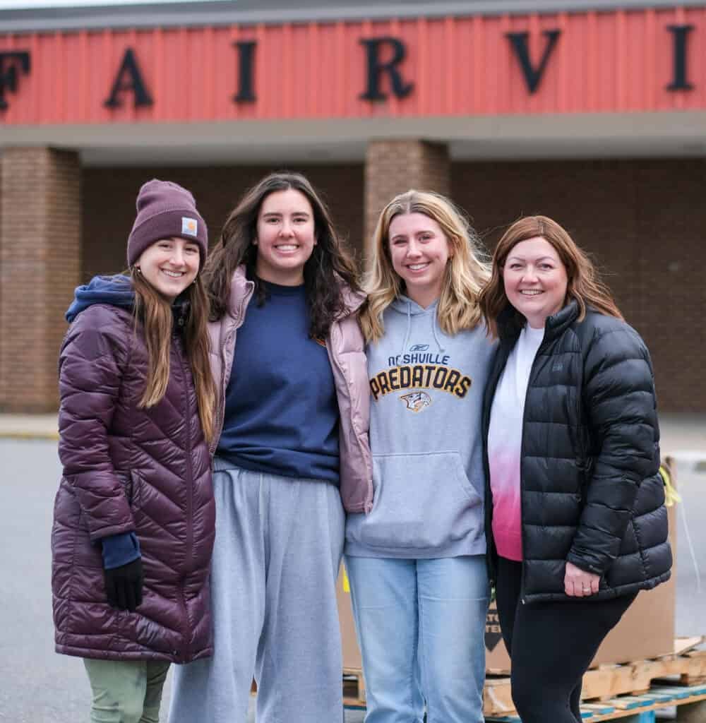 Four young women in winter jackets stand close together, smiling outside in front of a building with a red sign that reads FAIRV. Its cloudy and they appear happy and casual.