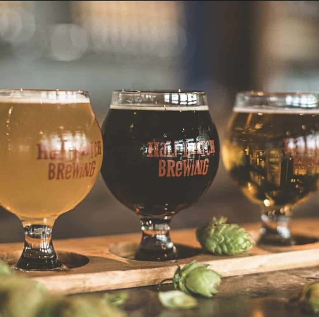 Three glasses of beer—light, dark, and golden—sit on a wooden paddle with hops scattered around. The glasses have Brewing printed on them and are on a blurred bar background.
