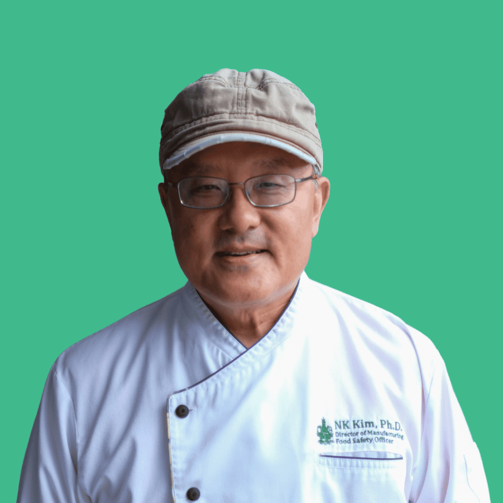 An older man wearing glasses, a light gray cap, and a white chef coat embroidered with N.K. Kim, Ph.D. stands smiling in front of a solid turquoise background.