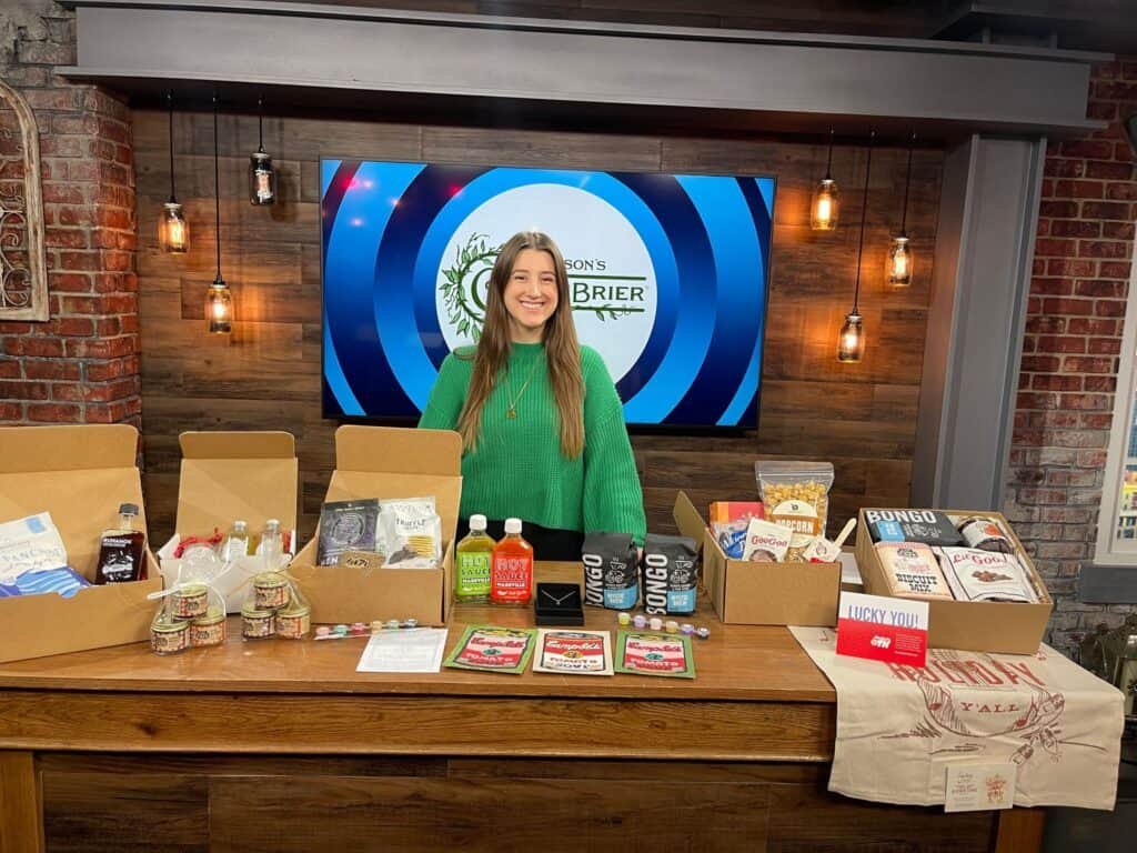 A smiling woman in a green sweater stands behind a wooden table displaying various snack boxes and drinks on a TV set. A screen behind her shows a circular logo with the text Greenbrier.