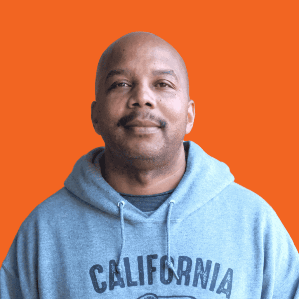 A man with a shaved head wearing a gray hoodie that says California stands in front of an orange background, looking slightly upward and smiling softly.