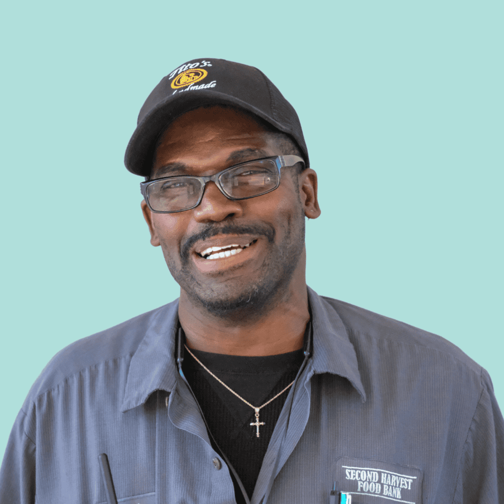 A smiling man wearing glasses, a black baseball cap, and a gray work shirt with a Second Harvest Food Bank patch stands against a light blue background.