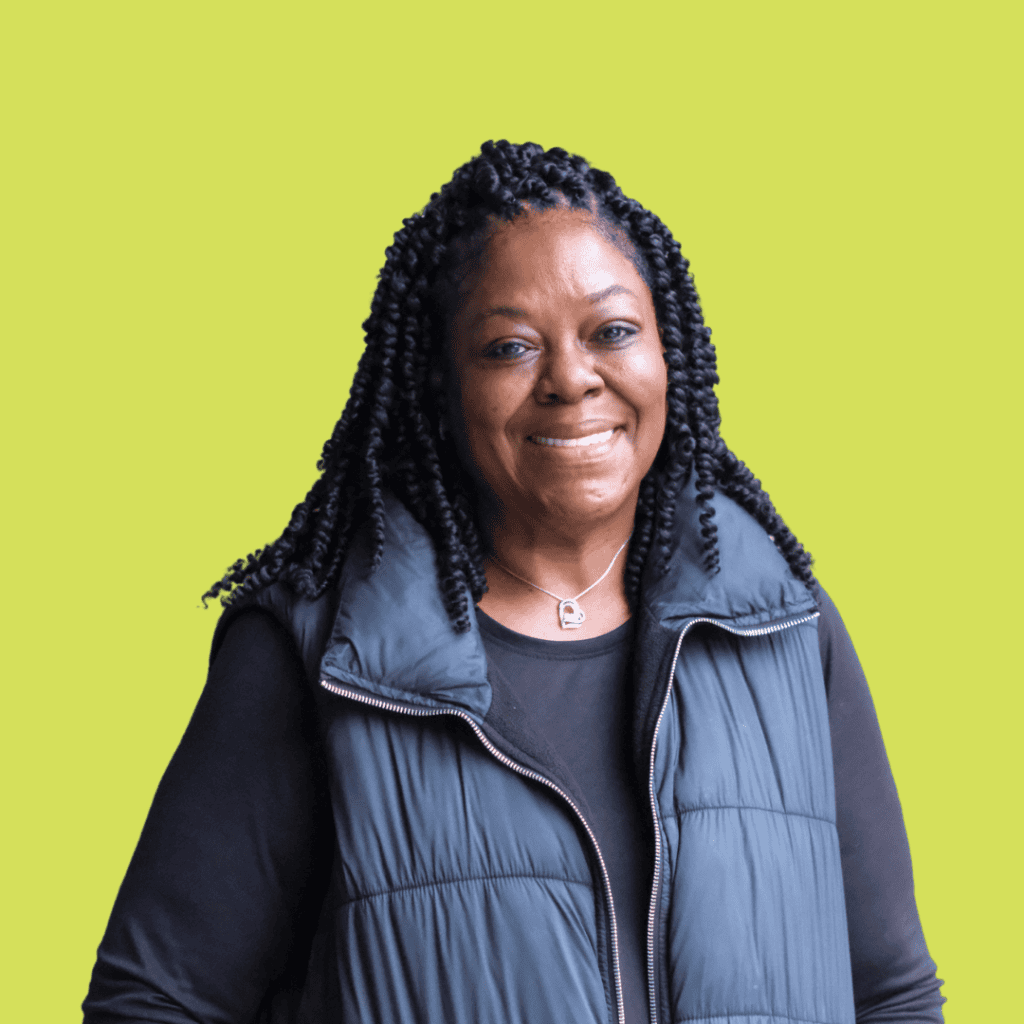 A smiling woman with long braided hair wearing a black shirt and a puffy vest stands in front of a bright yellow-green background.