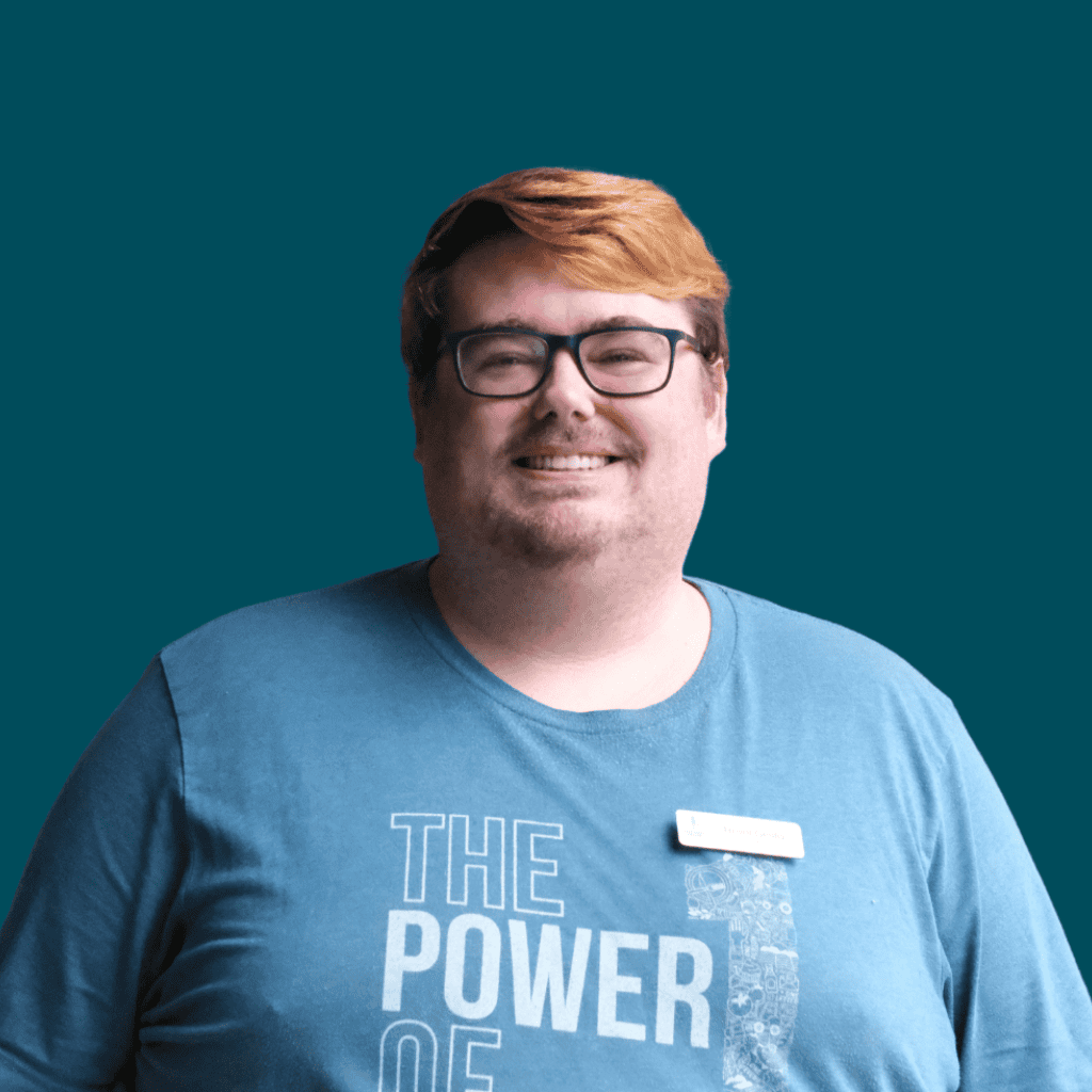 A smiling person with short, reddish-brown hair and glasses wearing a blue shirt that says THE POWER OF and a name badge, standing in front of a solid teal background.