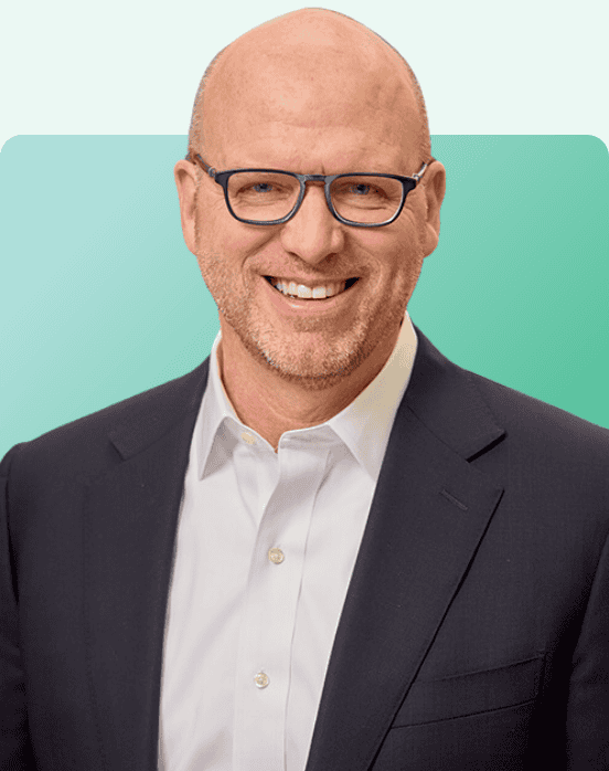 A smiling bald man wearing glasses, a white dress shirt, and a dark suit jacket, posing in front of a gradient turquoise background.