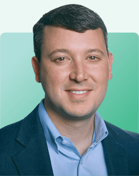 A man with short dark hair wearing a blue blazer and light blue shirt, smiling at the camera. The background is a gradient of light green shades.