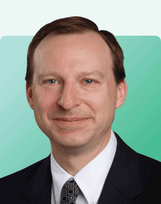 Professional headshot of a man with short light brown hair, wearing a dark suit, white shirt, and patterned tie, smiling slightly against a green and light blue gradient background.