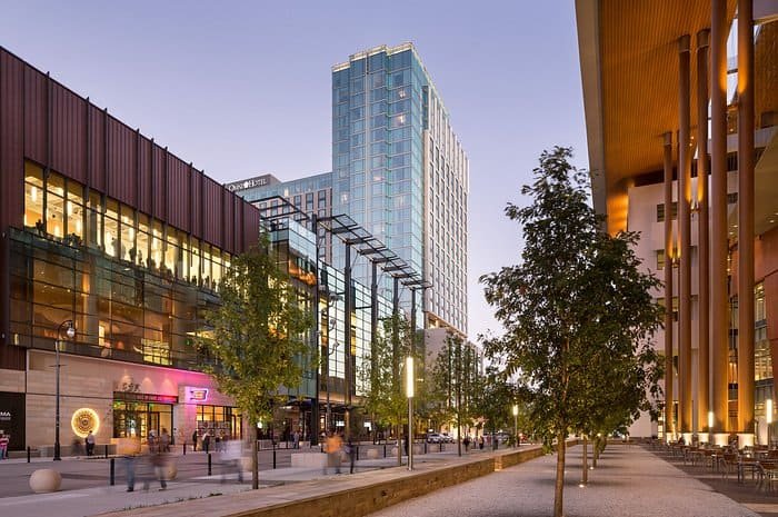 A modern city street at dusk with tall buildings, glass windows, bright lights, trees lining the sidewalk, and people walking along the urban outdoor area.