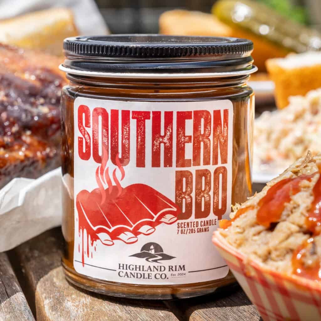 A jar candle labeled Southern BBQ Scented Candle by Highland Rim Candle Co. sits on a wooden table surrounded by barbecue food, including ribs, cornbread, pickles, and a dish with barbecue sauce.