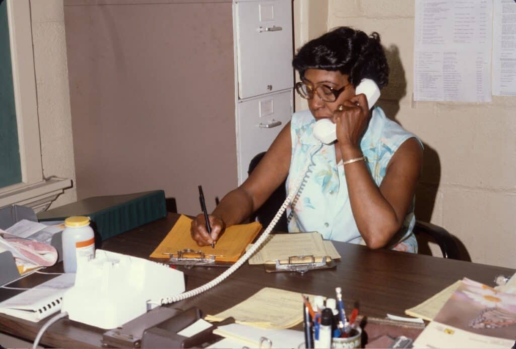 A woman wearing glasses sits at a desk, talking on a corded phone and writing on papers. The desk is cluttered with documents, a clipboard, a pill bottle, and office supplies. A filing cabinet and papers are in the background.