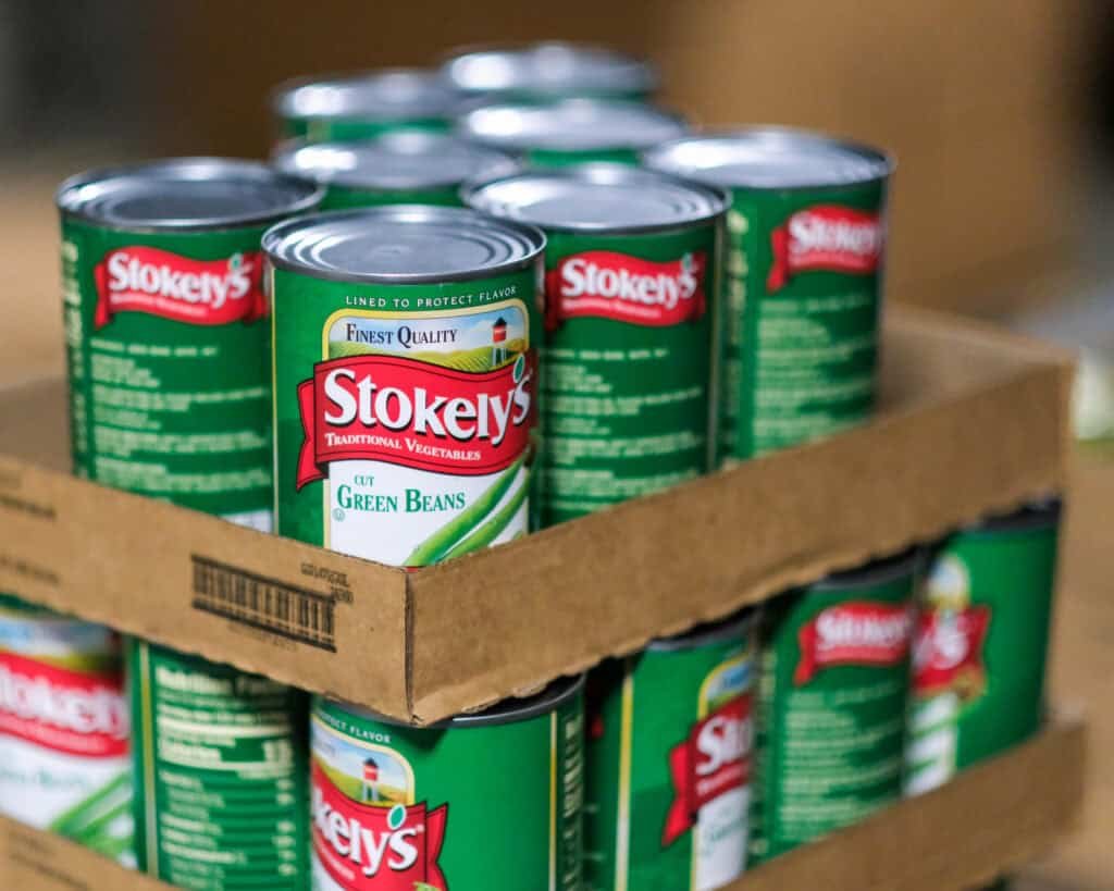 Several cans of Stokely’s cut green beans are stacked in a cardboard tray, with the labels facing forward. The cans are arranged in rows, and the background is blurred.