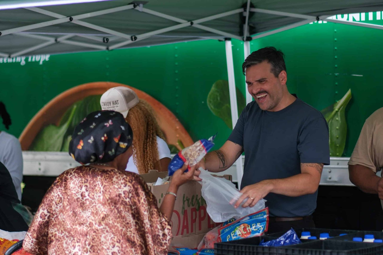 A smiling man hands a bag of food to a woman wearing a headscarf at an outdoor market or food distribution event, both under a tent with other people and bags of groceries in the background.