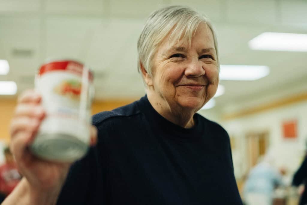 An older woman with short gray hair smiles at the camera while holding up a can of food in a brightly lit indoor setting.