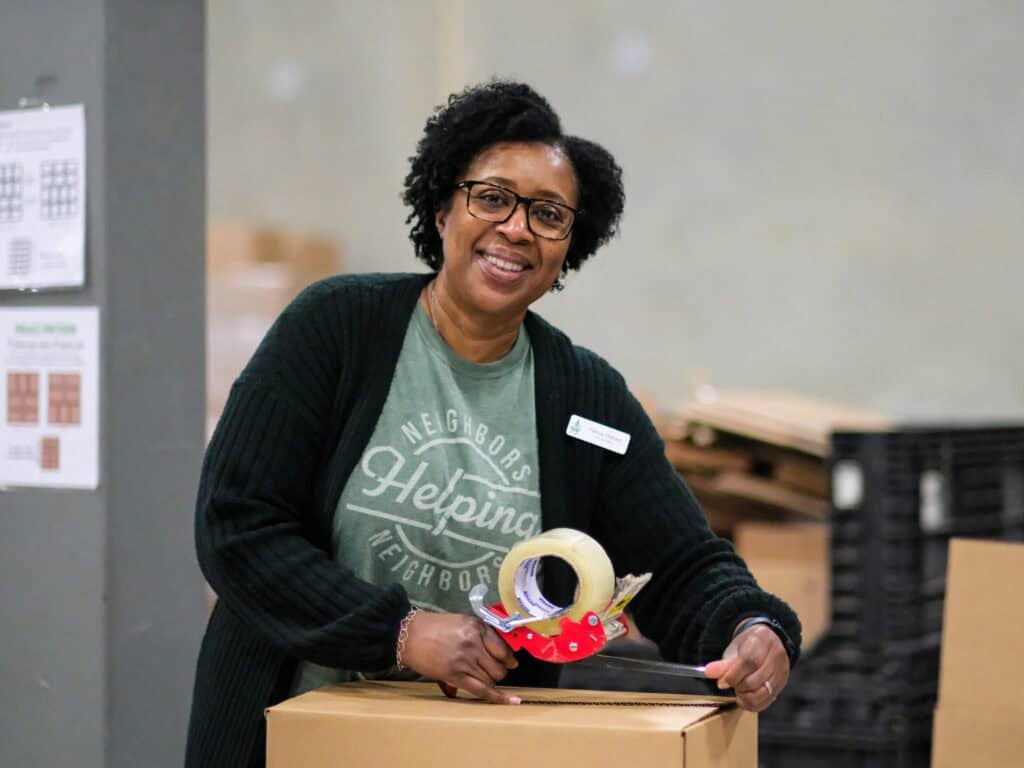 A smiling woman wearing glasses and a “Neighbors Helping Neighbors” shirt seals a cardboard box with packing tape in a warehouse setting.